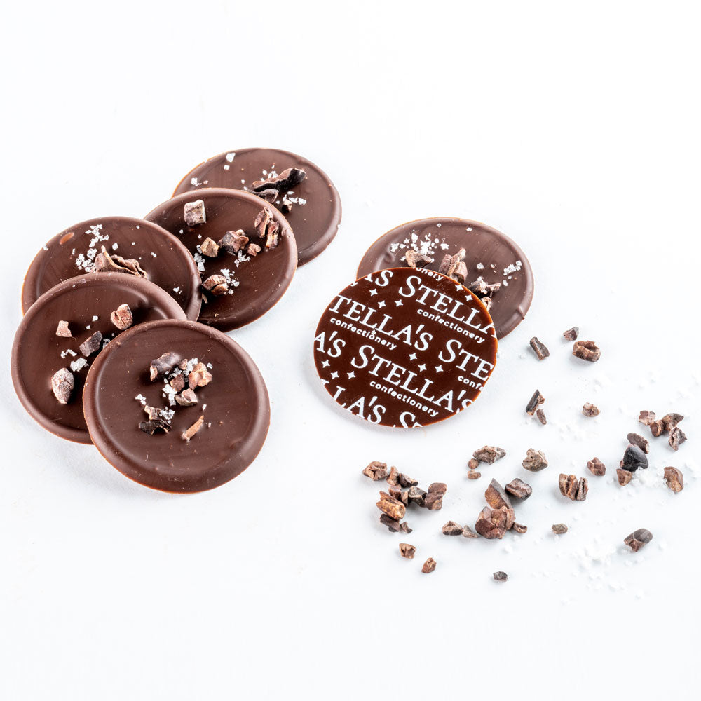 Chocolate discs stamped with Stella's Confectionery logo, surrounded by cocoa nibs and rock salt